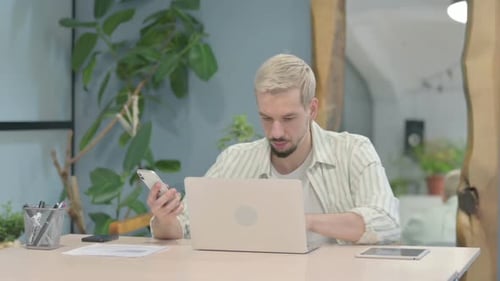 Man Types on Laptop While Looking at Phone