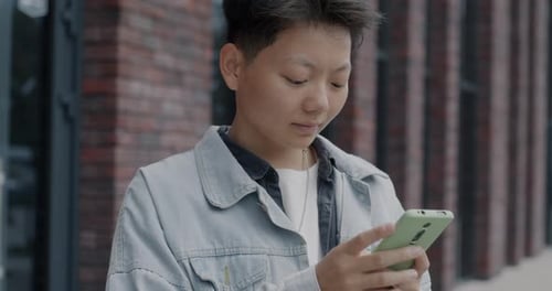 Slow Motion Portrait of Cheerful Asian Man Using Smartphone and Smiling Standing Outdoors in City