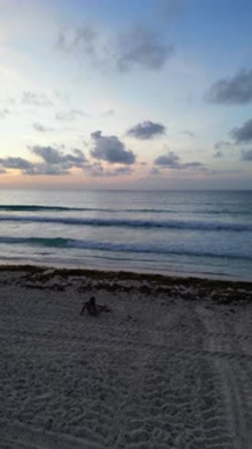 Aerial view of a woman sitting on the sand, with her back turned, watching the sunrise on a beach.