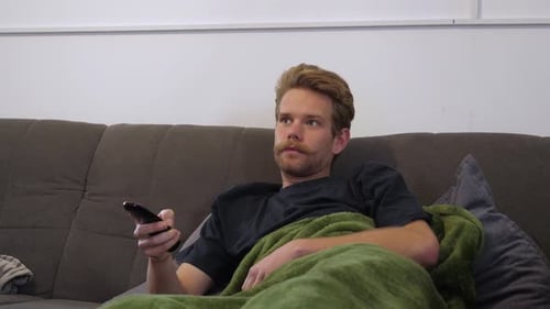 The blond handsome guy, with a lovely mustache lying on the couch while hes wrapped in his green flu