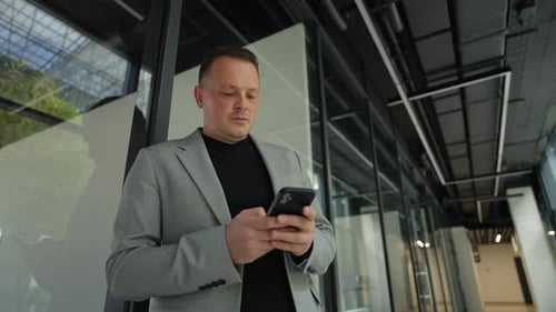 A Businessman in the Corridor of an Office Premises Sends an SMS on a Mobile Phone