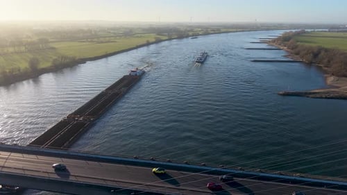 Aerial view of a wide river crossed by a busy bridge with cars, while a cargo barge and a passenger