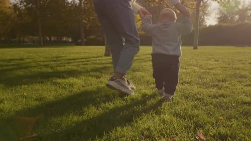 Mother guides young boy taking first steps on green lawn in park