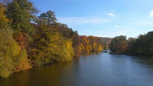 Beautiful autumn scenery. Calm blue river surrounded with yellow, red and orange tree under blue sky