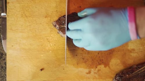 Top view, slo-mo footage, of a man wearing gloves slicing cooked, juicy, strips of steak on a wooden