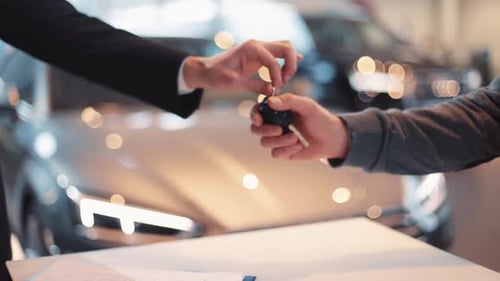 Young Man Giving Keys of Car to Buyer Men Shaking Hands in Beautiful Car Dealership on Background of