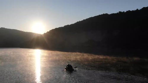 Aerial View of Two People in Silhouette Rowing a Boat in a Calm Misty River Surrounded by Hills Duri
