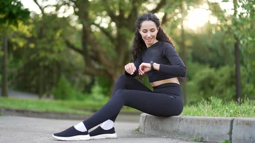 Smiling young sportswoman looking smart watch sitting in urban city park. Woman using checking