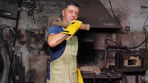 Blacksmith in Safety Apron Standing in Workshop with Huge Hammer on His Shoulder While Looking at