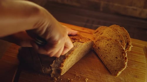 Close-up hands of female baker with knife cutting freshly baked aromatic loaf of wholemeal bread