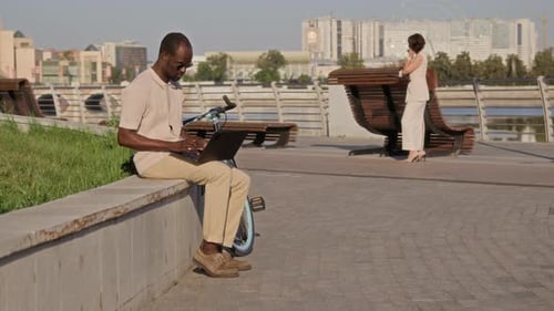 Young Man with Sunglasses Working on Laptop Sitting on Concrete Ledge near River