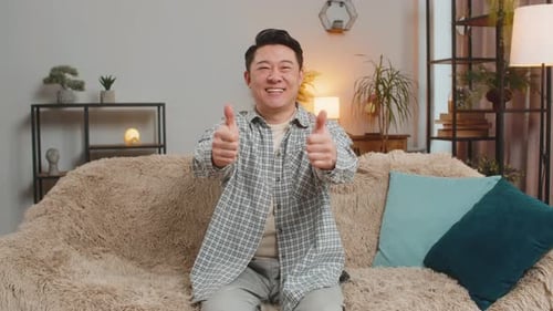 Portrait of Excited Smiling Chinese Young Man Showing Double Thumbs Up Sitting on Sofa Couch at Home