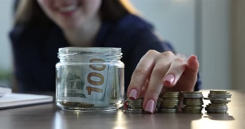 Woman Putting Coins into Savings Jar on Desk