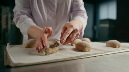 Bäckerin schneidet mit einem professionellen Bäckermesser Muster auf rohe Brötchen, bevor sie eine Bäckerei backt