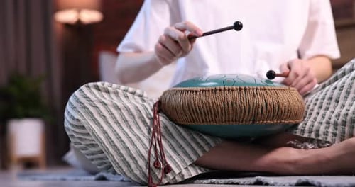 Woman playing steel tongue drum with mallets on floor at home, closeup