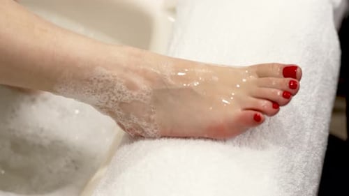 A woman takes her foot out of a bath with soapy water and places it on a pedicure stand, red gel pol