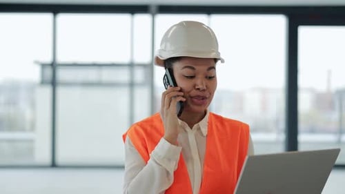 Engenheira confiante com capacete e colete segura laptop e fala ao telefone em novo escritório