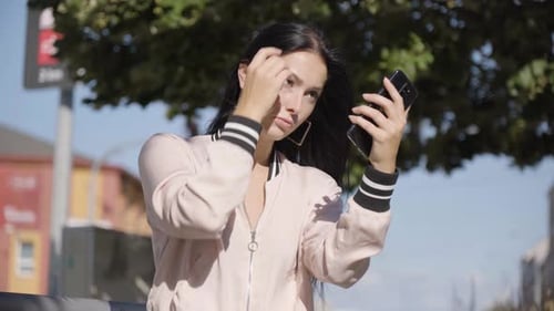 A Young Caucasian Woman Checks Herself and Fixes Herself on Her Smartphone in an Urban Area