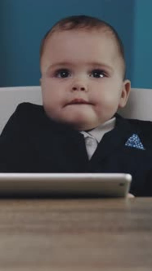 Baby Sits at Table in Suit by Tablet