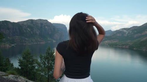 From a high vantage point, a woman with long dark hair stands by a fjord, gesturing towards the sere