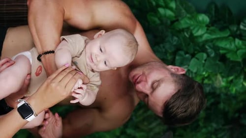 Caucasian dad holding a baby boy in front of him. Female hands clap tiny kid’s hands. Vertical view.