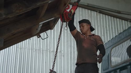 Schmutziger Mechaniker, asiatischer Junge, der in einer kleinen vietnamesischen Werkstatt einen Motor mit einem manuellen Hebezeug absenkt