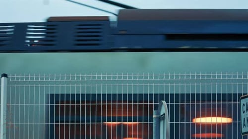 A fast passenger train rushing past behind a metal fence, with blurred motion and glowing interior l