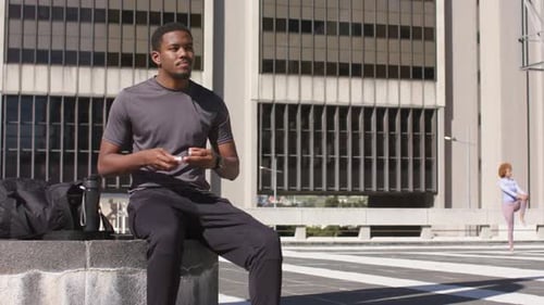 African American man opening earbud case on bench in sunlit plaza inserting earbuds for listening