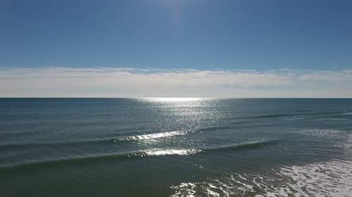 Drone shot moving away from the ocean's gentle waves, towards the beach. The sun reflects off the oc