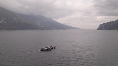 Droneshot of a ferry boat going to the city Limone Italy on Lake Garda on a grey day with mountains