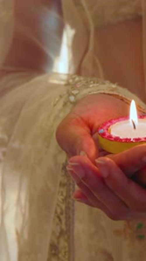 Vídeo vertical: mulher desenhando chamas em casa usando sari creme, segurando diya com borda rosa para puja