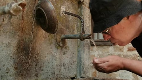 Man wearing hat and glasses cupping hands and drinking cold water to quench thirst from an old villa