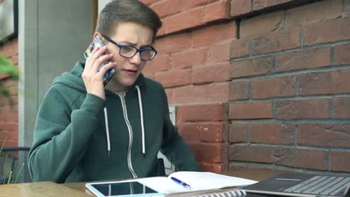 Young Teenager with Cellphone, Laptop and Notepad Doing Homework in Cafe Alone