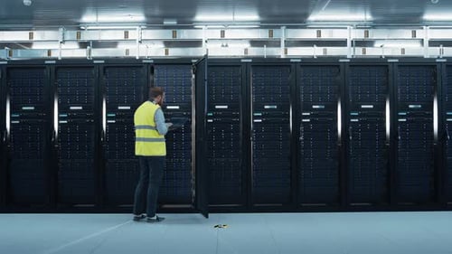 IT Specialist in High Visibility Vest is Working on Laptop in Data Center while Standing Before Ser
