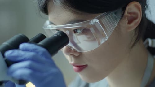 Asiatische Wissenschaftlerin mit Schutzbrille schaut durchs Mikroskop
