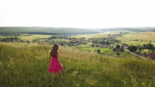 Yong Woman with Long Hair in Red Dress Walking in Summer Field with Tall Green Grass on Rural Hill