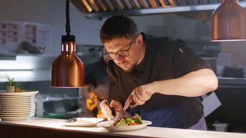 Chef Plating Gourmet Dish with Tweezers under Heat Lamp in Restaurant Kitchen
