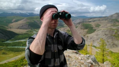 Handsome Gay Explorer Standing Proudly on a Mountain Peak Looks Through Binoculars Surrounded By a