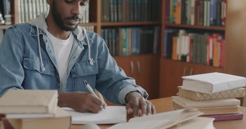 African American Man Writing Information in Notebook Studying Books in Library Getting Knowledge