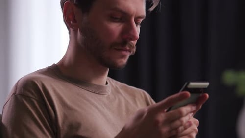 A Young Man with Stubble Uses a Smartphone A Man in a Brown Tshirt is Holding a Phone and Typing a