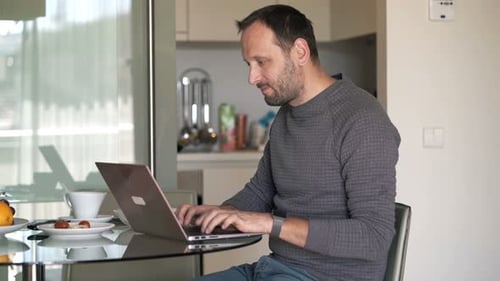 Man Working On Laptop Sitting By Table In Kitchen At Home