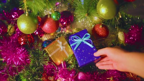 An Unrecognizable Hand Puts Blue Box with Gift Next to Yellow One They Lies on Colorful Tinsel
