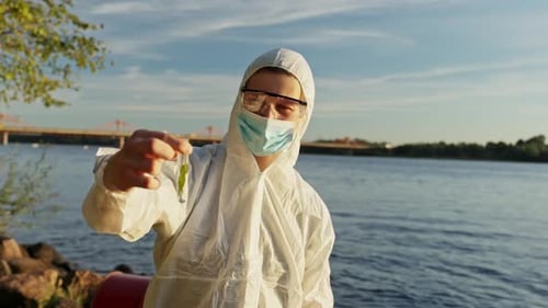 Young caucasian environmental inspector in hazmat suit collecting water samples from polluted river