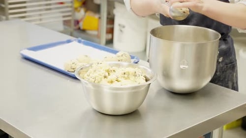 Woman Scoops Cookie Dough from bowl on Stainless Steel Table in Professional Kitchen