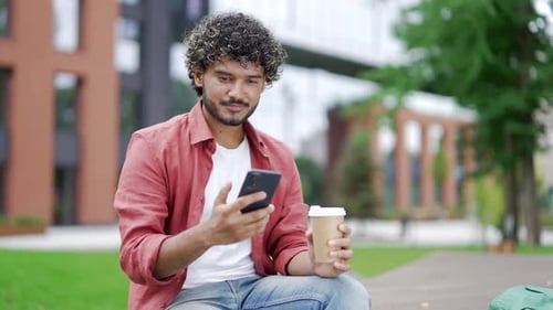 Young man uses a mobile phone while sitting on a bench on street near office building. Handsome male