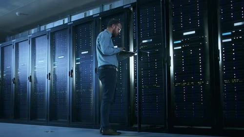 Bearded IT Specialist in Glasses is Working on Laptop in Data Center while Standing Before Server R