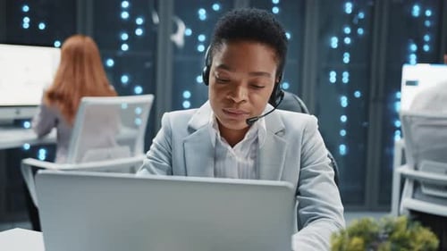 Ernsthafte Afroamerikanerin mit Headset trägt formelle Kleidung beim Sitzen, benutzt Laptop, arbeitet mit