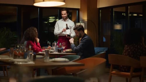 Waiter Pouring Wine Bottle to Fancy Couple Table at Night Restaurant Dinner