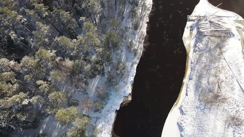 Aerial view of a winding river through a dense snow-covered forest in winter, with a curving road ru