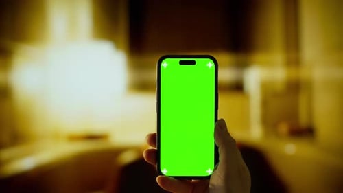 Person Holding Smartphone with Bright Green Screen in Dimly Lit Indoor Room Showcasing Mobile